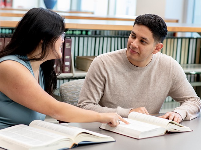 Two students studying in the library with a book at a desk