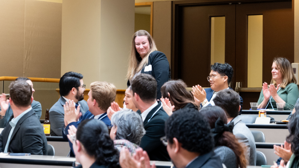 A group of people seated at tables lined up in rows applaud a person standing in the back of the room