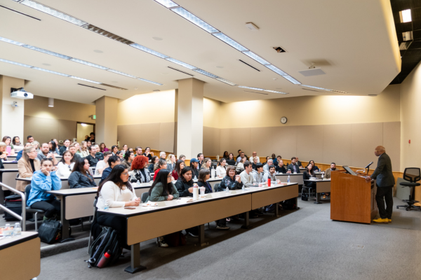 A full lecture hall listens to Professor Kennedy deliver a lecture at the front behind a wooden lectern.