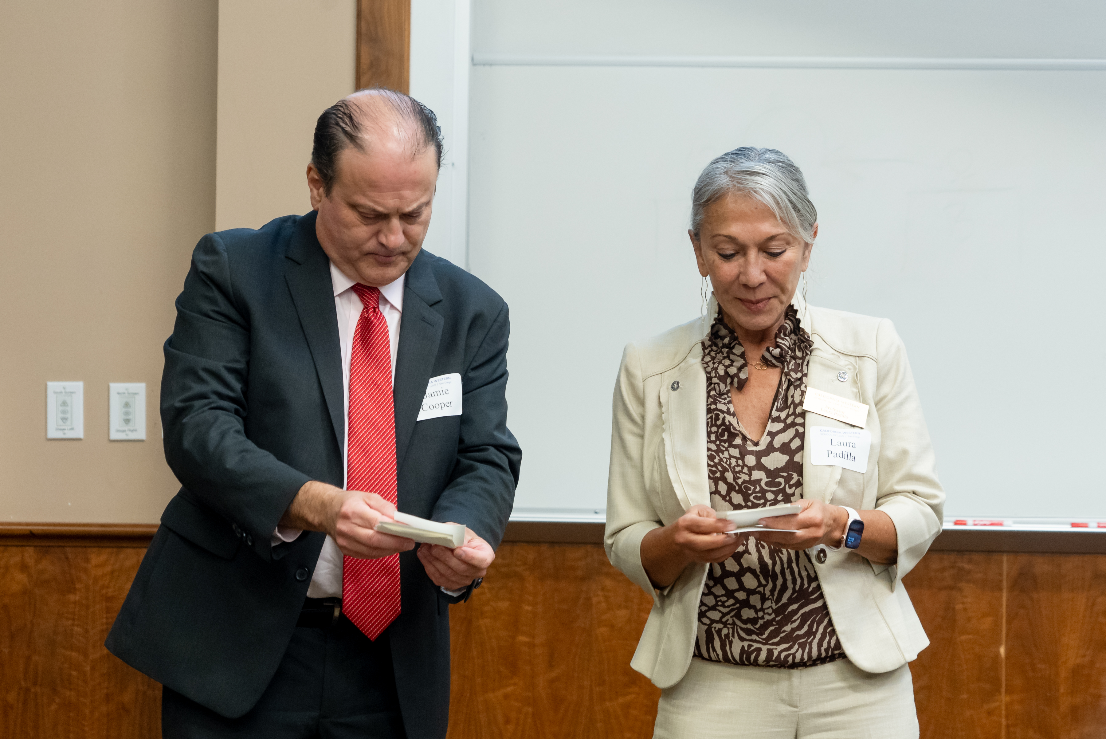 Two individuals, a man in a dark suit with a red tie and a woman in a light suit, are standing and reading from notecards in a classroom.