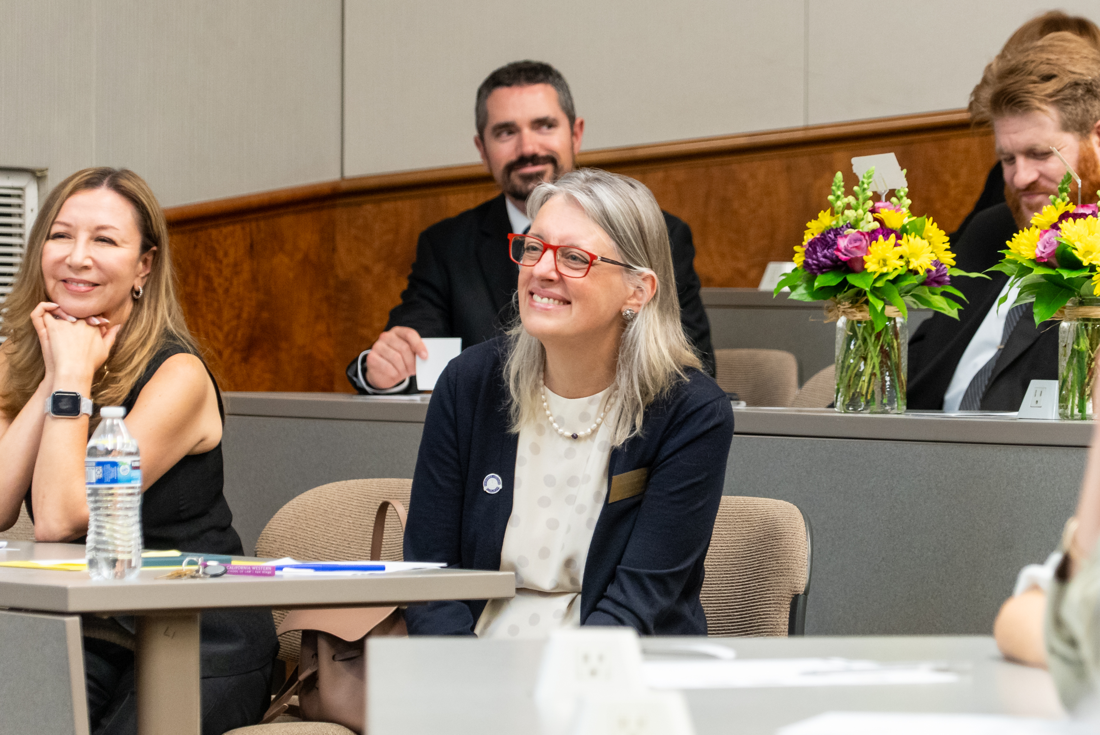 People seated in a classroom, smiling and engaged; two vases of colorful flowers are on desks in the background.